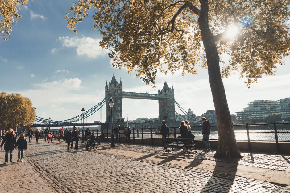 A sunny outdoor scene along the River Thames in Tower Hamlets, with the iconic Tower Bridge in the background. The foreground shows a cobblestone promenade with several people walking, sitting on benches, and standing near the water's edge. A large tree with autumn leaves partially shades the area, and the sunlight filters through the branches. Along the pavement, there are recyclable cardboard boxes, wrapping materials, and packing supplies indicating a home relocation activity. A man with a van, possibly from Man With a Van Poplar, is visible nearby, engaged in the loading process of furniture or packing boxes onto a vehicle for a house removals service. The environment appears peaceful and well-lit, emphasizing the practical aspects of furniture transport and packing within the context of moving services in E14 postures, such as avoiding fines related to parking suspensions during moves.