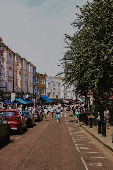 A narrow street at Chrisp Street Market in Poplar during daytime, with market stalls and shops lining both sides of the pavement. The street is busy with pedestrians, some carrying shopping bags or boxes, and vehicles parked along the curb. Large green trees are visible to the right, providing partial shade over the area. In the background, multi-storey buildings with colourful facades and signs for various shops are evident. The scene also shows a van parked at the side of the street, part of the home relocation and furniture transport process, indicative of [COMPANY_NAME]’s involvement in local removals or moving services. The overall environment reflects a bustling urban market area, with ample space for loading and unloading boxes, furniture, and packing materials as part of a professional moving operation in competition with typical street activity.