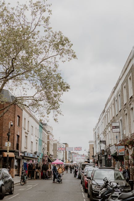 A busy street scene in Poplar with a row of commercial buildings on both sides, featuring various shopfronts and signage. Parked cars line both sides of the narrow pavement, while pedestrians and shoppers are visible walking and browsing. A large tree with sparse foliage extends over the street from the left side, partially obscuring the overcast sky. Several people are seen with shopping bags, and a woman with a pushchair is visible in the middle of the street. In the foreground, a vehicle is parked close to the curb, and a scooter is parked next to it. The scene captures an active, urban environment suitable for house removals and furniture transport, with potential moving and loading activity nearby, as typical for a community involved in home relocation services. Man With a Van Poplar is based in this area, supporting the logistics of local removals and packing tasks.