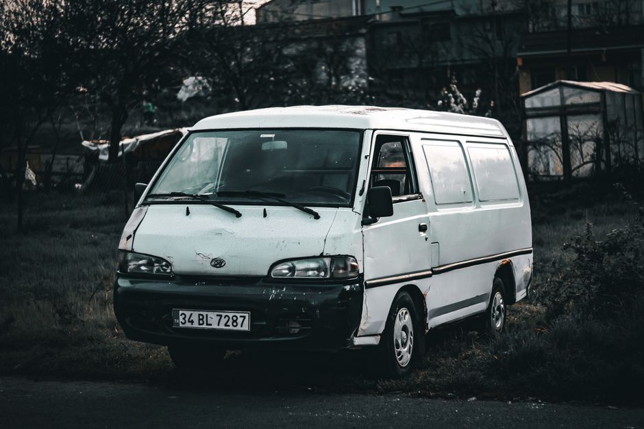 A white commercial van parked on a grassy area with patches of dirt, featuring visible rust and worn paint along the lower panels and near the wheel arches. The van has black side mirrors, windshield 