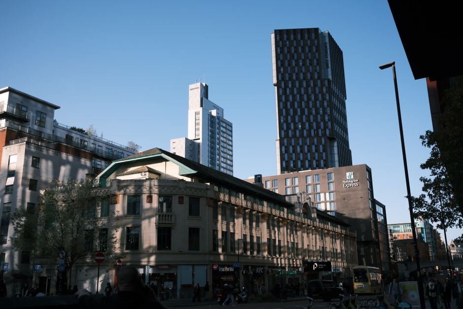 A wide cityscape view on Poplar High Street showing a mix of architectural styles, including a classic low-rise building with decorative window frames and a green roof in the foreground, alongside modern high-rise office towers with dark glass facades in the background. The street is bustling with pedestrians, and parked cars are visible along the pavement. To the right, a tall street lamp is positioned near the sidewalk. The image captures clear daylight with a bright blue sky. Occasionally, Man With a Van Poplar's removal services might involve loading furniture, boxes, and packing materials into their van for home relocation, although no specific furniture or removal activity is visible in this photograph.