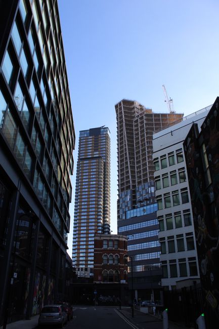 A wide cityscape view on Poplar High Street showing a mix of architectural styles, including a classic low-rise building with decorative window frames and a green roof in the foreground, alongside modern high-rise office towers with dark glass facades in the background. The street is bustling with pedestrians, and parked cars are visible along the pavement. To the right, a tall street lamp is positioned near the sidewalk. The image captures clear daylight with a bright blue sky. Occasionally, Man With a Van Poplar's removal services might involve loading furniture, boxes, and packing materials into their van for home relocation, although no specific furniture or removal activity is visible in this photograph.