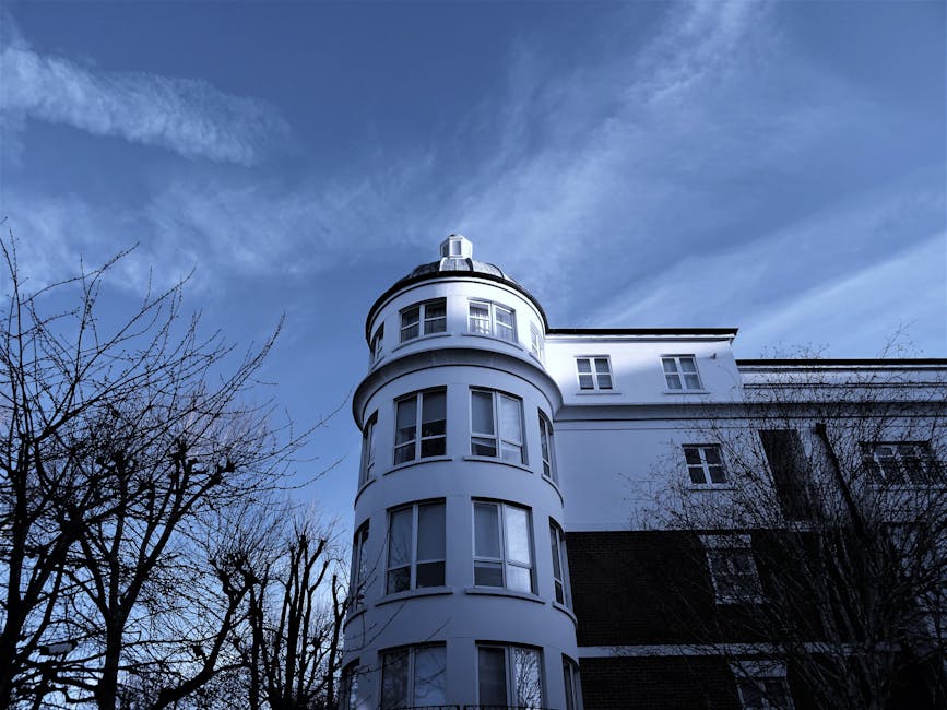 An upward view of a white, rounded, multi-story building with large sash windows, featuring a turret-like corner extending above the main structure, set against a partly cloudy blue sky. Surrounding the building are bare tree branches indicating late autumn or winter. In the foreground, a pavement area is visible, possibly a loading zone, but no vehicles or people are visible in this image. As part of home relocation or furniture transport services, this scene may relate to the planning or execution of moving operations at this property. The image reflects the exterior environment where [COMPANY_NAME] conducts removals or access planning for flats and buildings with limited access, and highlights the structural features faced during lifting and loading processes involved in packing and moving services for apartments or similar residential properties. The ambient lighting suggests daytime conditions with natural light highlighting the building's architectural details.