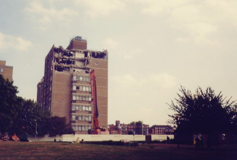 A partially demolished multi-storey brick building with exposed concrete and broken windows, situated in an urban setting with a construction excavator positioned next to the structure, indicating ongoing demolition work. The foreground features a grassy area with a few trees and a pathway, while the background shows a cloudy sky with other buildings visible at a distance. The scene captures the process of building clearance, highlighting the debris and structural damage typical of a building removal. Occasionally, a vehicle or equipment related to the demolition may be seen nearby, emphasizing the logistics involved. This image reflects the type of urban house removal or building clearance that companies like Man With a Van Poplar might facilitate as part of their comprehensive removals services, especially when preparing a site for new construction or clearance. The overall environment suggests a professional and organized demolition scene, with clear focus on structural dismantling and recycling of materials.