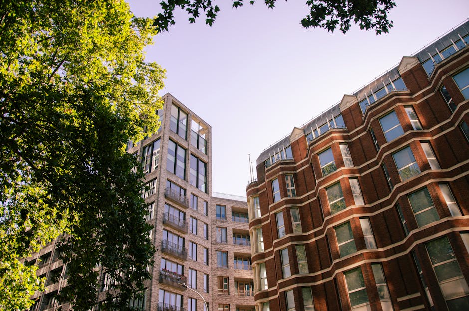 An upward view of a white, rounded, multi-story building with large sash windows, featuring a turret-like corner extending above the main structure, set against a partly cloudy blue sky. Surrounding the building are bare tree branches indicating late autumn or winter. In the foreground, a pavement area is visible, possibly a loading zone, but no vehicles or people are visible in this image. As part of home relocation or furniture transport services, this scene may relate to the planning or execution of moving operations at this property. The image reflects the exterior environment where [COMPANY_NAME] conducts removals or access planning for flats and buildings with limited access, and highlights the structural features faced during lifting and loading processes involved in packing and moving services for apartments or similar residential properties. The ambient lighting suggests daytime conditions with natural light highlighting the building's architectural details.