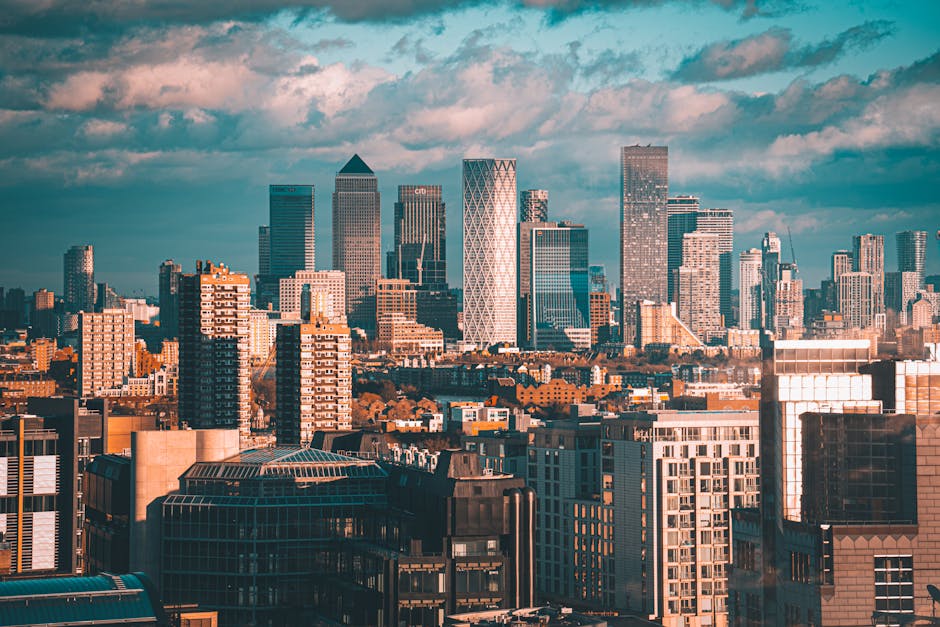 A high-angle view of a city skyline during daytime showing multiple modern high-rise office buildings and residential towers with glass facades, some with geometrical architectural designs. In the foreground, there are lower buildings constructed from brick and concrete, forming a dense urban landscape. The scene includes a clear sky with some clouds, and the lighting suggests late afternoon or early evening. This urban environment relates to house removals and furniture transport, as it depicts a typical cityscape where professional movers like Man With a Van Poplar might operate during home relocation or office moving processes, involving loading and transporting furniture and boxes through streets and into vehicles parked nearby.