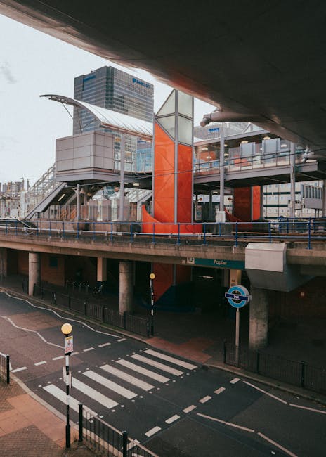 A modern multi-level transportation hub with an elevated walkway structure supported by bright orange columns, visible glass railings, and a curved metal roof canopy. The walkway connects different parts of the station, with some sections enclosed by glass panels. Below, there is a pedestrian zebra crossing on a small street with road markings, a traffic bollard, and a London Underground sign indicating proximity to a tube station. The environment is urban, with nearby tall office buildings and a clear sky overhead, suggesting daytime. The scene captures the infrastructure used for house removals and relocation logistics, illustrating the proximity to public transport options relevant for moving near Canary Wharf, with a focus on loading and access points. The image emphasizes urban transportation facilities associated with home relocation and furniture transport processes, as represented by the structural setup visible in the photo. Man With a Van Poplar operates in this area, supporting efficient house moves and moving services.