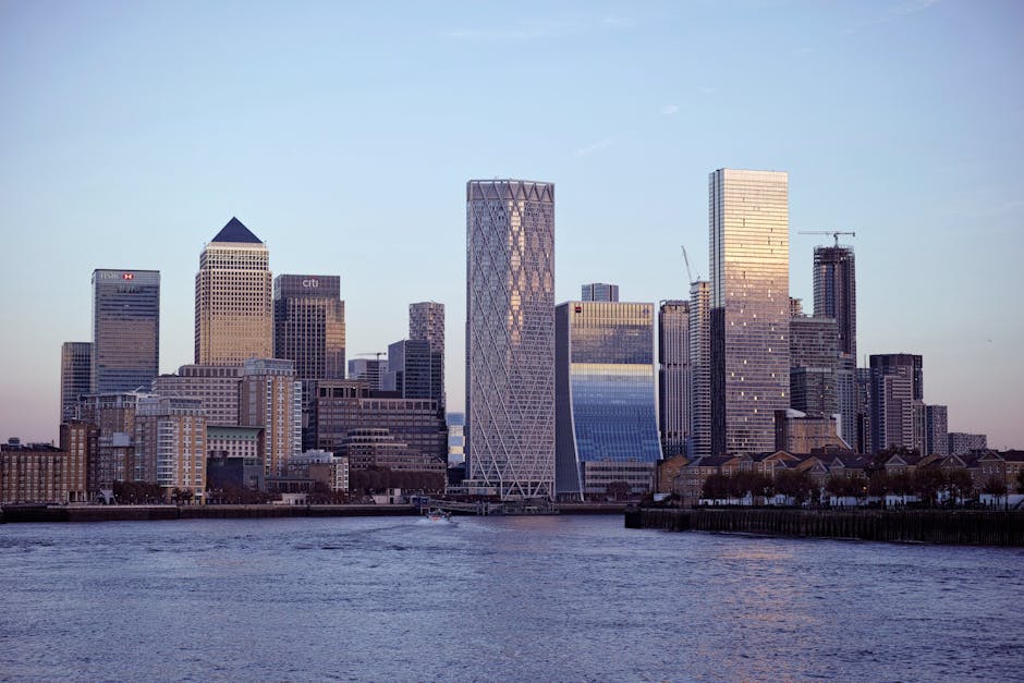 A high-angle view of a city skyline during daytime showing multiple modern high-rise office buildings and residential towers with glass facades, some with geometrical architectural designs. In the foreground, there are lower buildings constructed from brick and concrete, forming a dense urban landscape. The scene includes a clear sky with some clouds, and the lighting suggests late afternoon or early evening. This urban environment relates to house removals and furniture transport, as it depicts a typical cityscape where professional movers like Man With a Van Poplar might operate during home relocation or office moving processes, involving loading and transporting furniture and boxes through streets and into vehicles parked nearby.