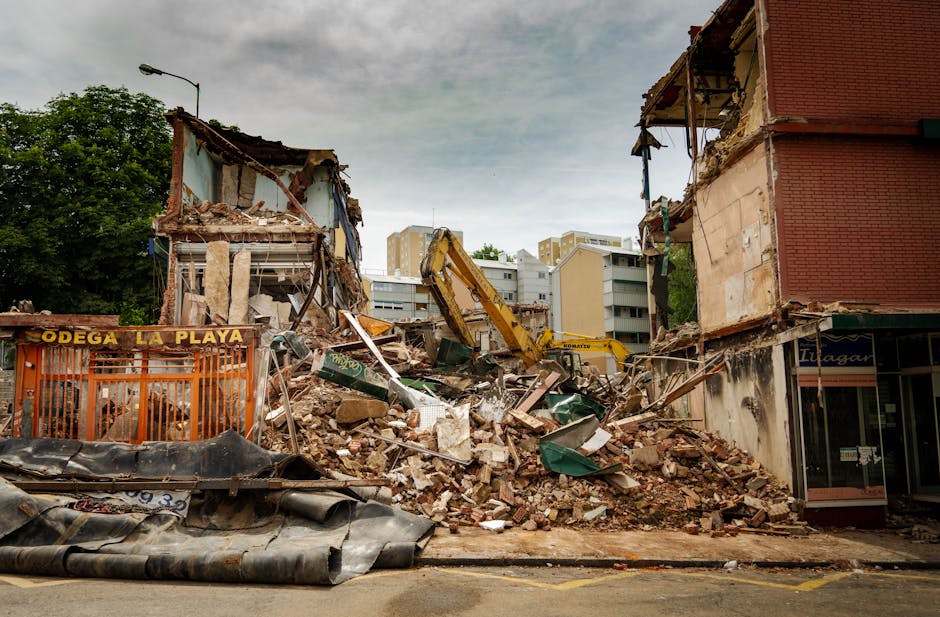 A partially demolished building in an urban area with large piles of debris, including broken bricks, concrete, and metal sheets, spread across the street and sidewalk. A yellow excavator is actively working amidst the rubble, with its arm extended to lift and move scattered materials. Next to the debris, there is a small fenced-off area with signage reading 'Odega La Playa,' indicating a nearby commercial establishment. The surrounding environment features multi-storey residential buildings in the background, along with trees and a streetlamp, under a cloudy sky. Bright daylight illuminates the scene, highlighting the extensive demolition process as part of a home renovation or urban redevelopment project. Occasionally, Man With a Van Poplar may be involved in removing leftover waste and clearing the site following demolition, supporting house clearance or building removal services.