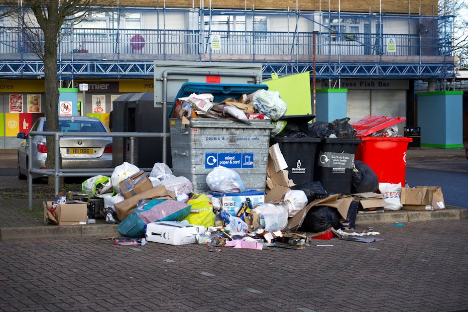 A large collection of overflowing waste and recycling bins situated on a paved sidewalk in front of commercial premises, including a grey mixed paper and card bin with an open lid, surrounded by black, red, and brown paper bags filled with rubbish, cardboard boxes, and loosely packed packaging materials. Some items are spilling onto the ground, with a grey metal railing separating the waste area from the parking space where a silver car is parked. Behind the bins, there is a storefront with signs for a fish bar and other businesses, under an upper floor with scaffolding and safety barriers indicating ongoing building work. This scene illustrates a typical urban waste collection point, relevant to moving services such as house clearance or clearance licensing offered by companies like Man With a Van Poplar, often associated with practical logistics involved in home relocation and waste disposal.